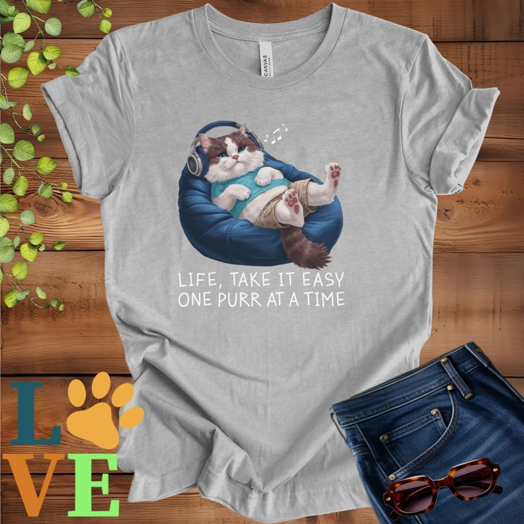 Light grey heather (athletic heather) t-shirt featuring an anthropomorphic Ragdoll cat relaxing on a bean bag while listening to music through headphones, with the slogan "Life, take it easy. One purr at a time."