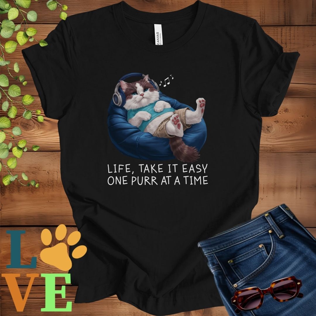 Black t-shirt featuring an anthropomorphic Ragdoll cat relaxing on a bean bag while listening to music through headphones, with the slogan "Life, take it easy. One purr at a time."
