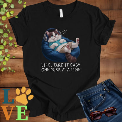 Black t-shirt featuring an anthropomorphic Ragdoll cat relaxing on a bean bag while listening to music through headphones, with the slogan "Life, take it easy. One purr at a time."
