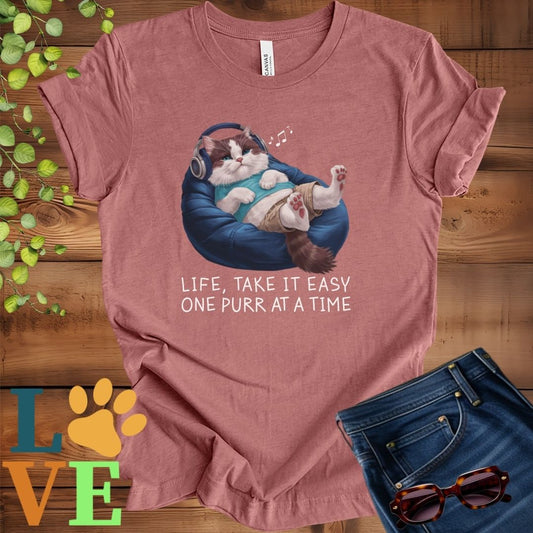 Muted mauve-purple (heather mauve) t-shirt featuring an anthropomorphic Ragdoll cat relaxing on a bean bag while listening to music through headphones, with the slogan "Life, take it easy. One purr at a time."
