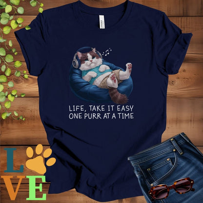 Navy blue t-shirt featuring an anthropomorphic Ragdoll cat relaxing on a bean bag while listening to music through headphones, with the slogan "Life, take it easy. One purr at a time."
