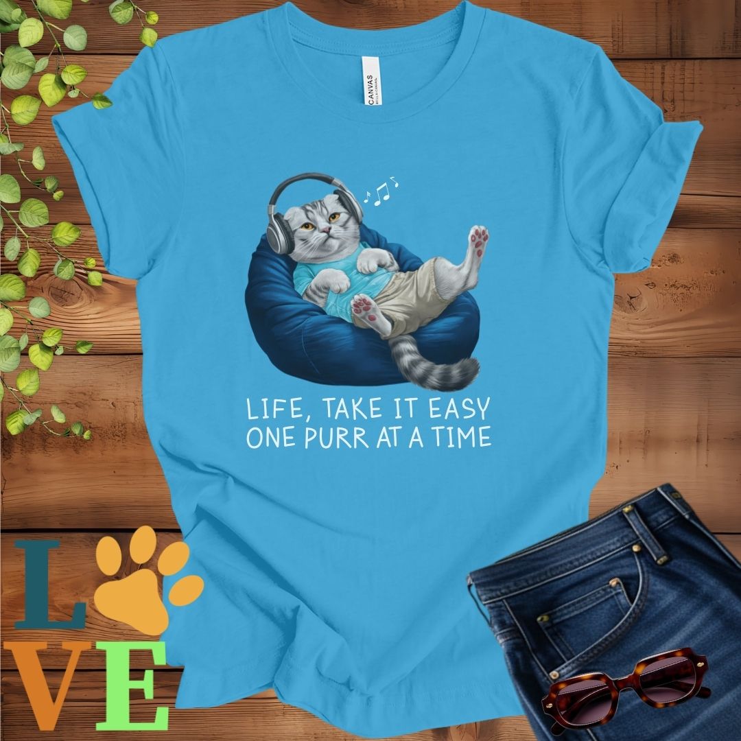Bright aqua blue t-shirt featuring an anthropomorphic Scottish Fold cat relaxing on a bean bag while listening to music through headphones, with the slogan "Life, take it easy. One purr at a time."
