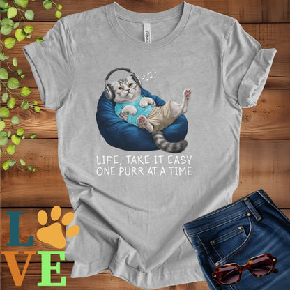 Light grey heather (athletic heather) t-shirt featuring an anthropomorphic Scottish Fold cat relaxing on a bean bag while listening to music through headphones, with the slogan "Life, take it easy. One purr at a time."
