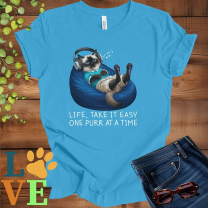 Bright aqua blue t-shirt featuring an anthropomorphic Siamese cat relaxing on a bean bag while listening to music through headphones, with the slogan "Life, take it easy. One purr at a time."
