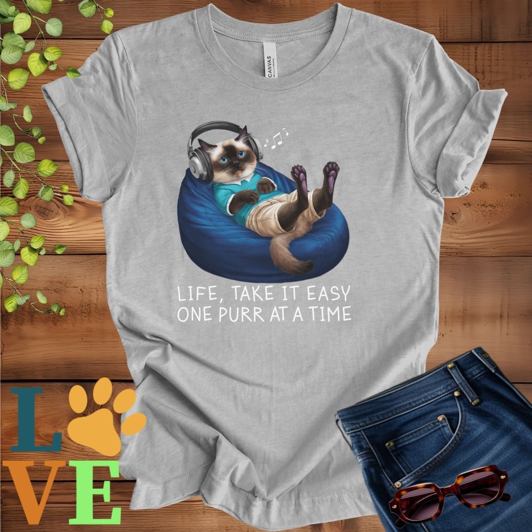 Light grey heather (athletic heather) t-shirt featuring an anthropomorphic Siamese cat relaxing on a bean bag while listening to music through headphones, with the slogan "Life, take it easy. One purr at a time."
