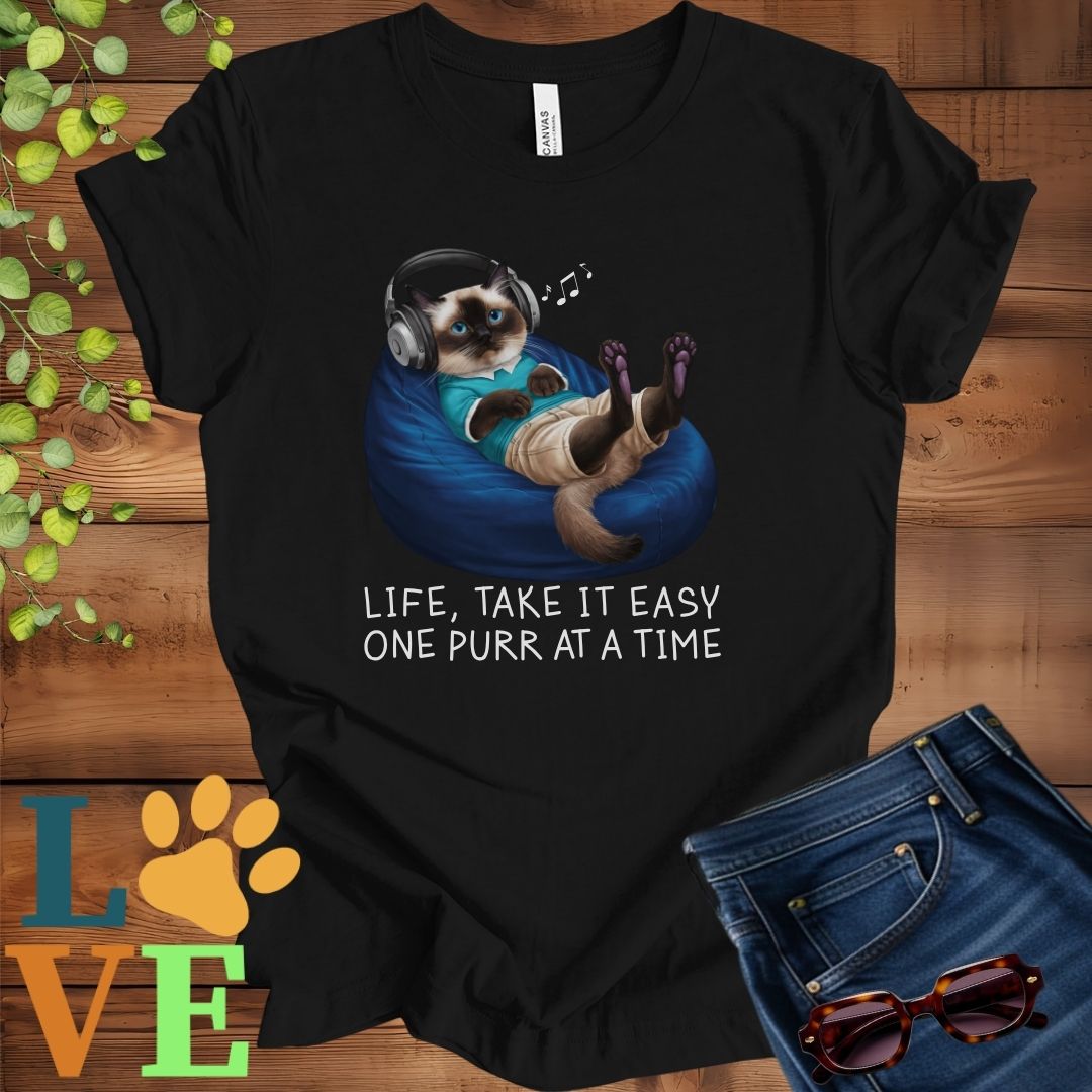 Black t-shirt featuring an anthropomorphic Siamese cat relaxing on a bean bag while listening to music through headphones, with the slogan "Life, take it easy. One purr at a time."
