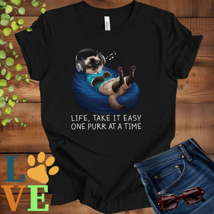 Black t-shirt featuring an anthropomorphic Siamese cat relaxing on a bean bag while listening to music through headphones, with the slogan "Life, take it easy. One purr at a time."

