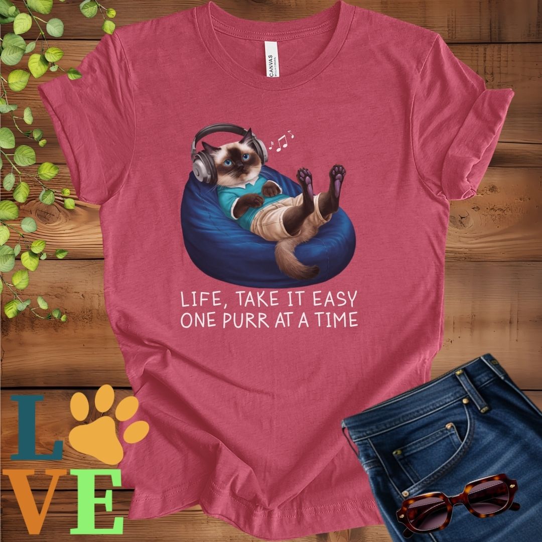 Muted raspberry pink (heather raspberry) t-shirt featuring an anthropomorphic Siamese cat relaxing on a bean bag while listening to music through headphones, with the slogan "Life, take it easy. One purr at a time."
