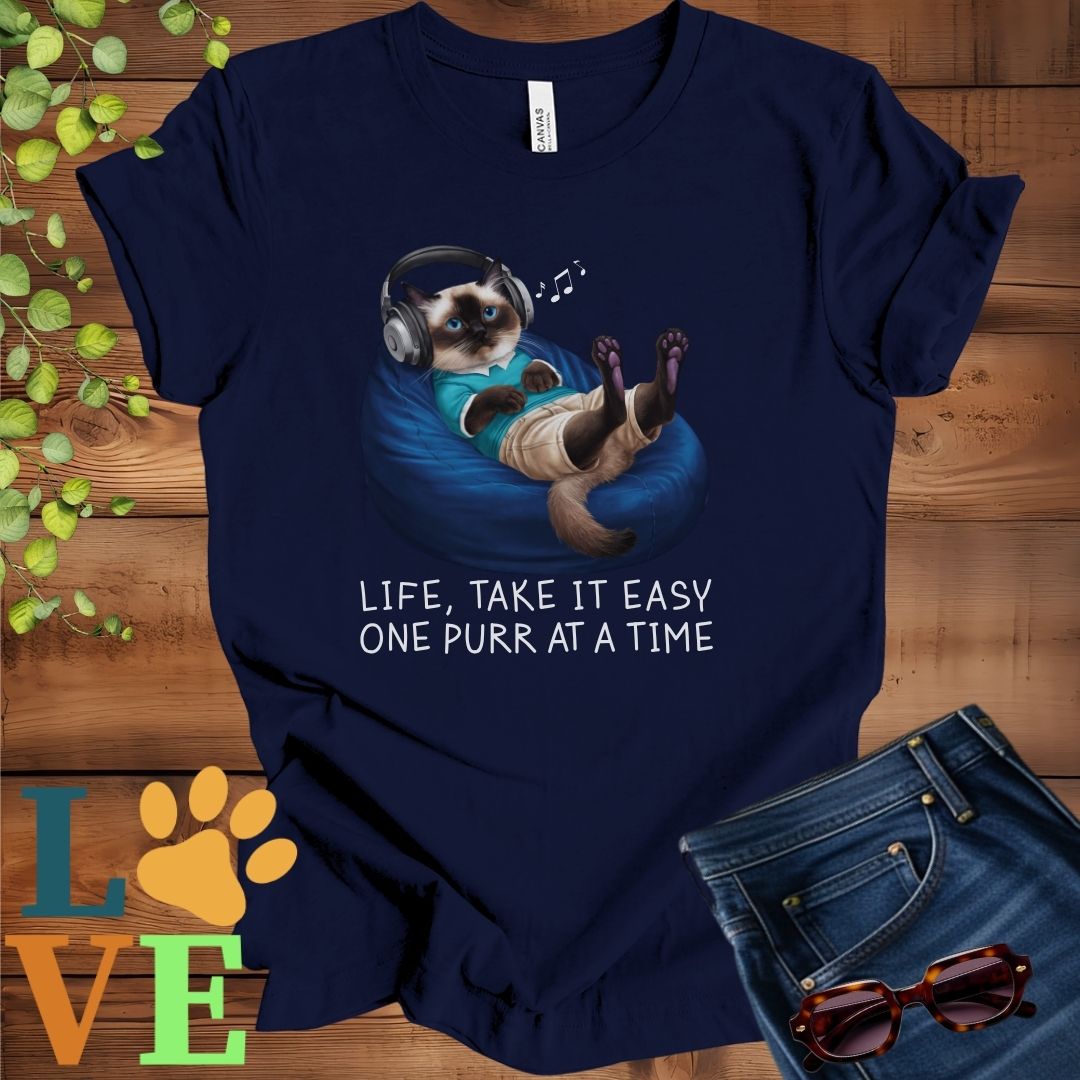 Navy blue t-shirt featuring an anthropomorphic Siamese cat relaxing on a bean bag while listening to music through headphones, with the slogan "Life, take it easy. One purr at a time."
