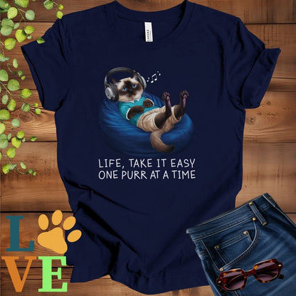 Navy blue t-shirt featuring an anthropomorphic Siamese cat relaxing on a bean bag while listening to music through headphones, with the slogan "Life, take it easy. One purr at a time."

