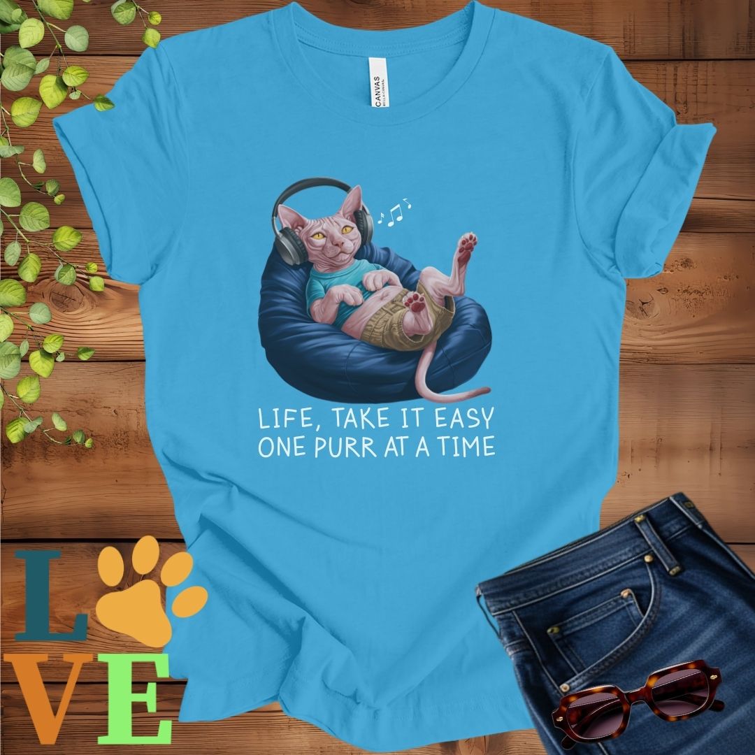 Bright aqua blue t-shirt featuring an anthropomorphic Sphynx cat relaxing on a bean bag while listening to music through headphones, with the slogan "Life, take it easy. One purr at a time."