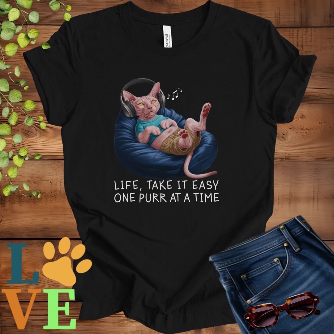 Black t-shirt featuring an anthropomorphic Sphynx cat relaxing on a bean bag while listening to music through headphones, with the slogan "Life, take it easy. One purr at a time."
