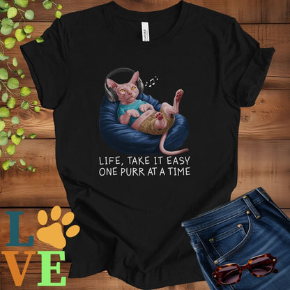 Black t-shirt featuring an anthropomorphic Sphynx cat relaxing on a bean bag while listening to music through headphones, with the slogan "Life, take it easy. One purr at a time."