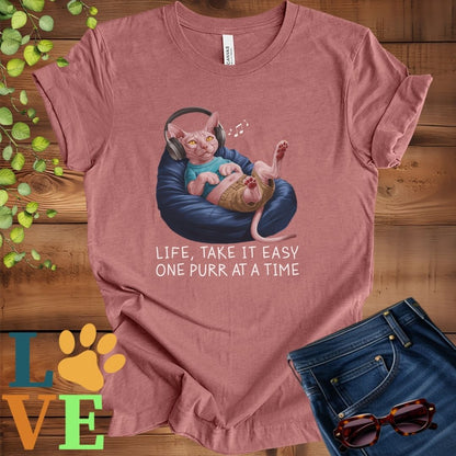 Muted mauve-purple (heather mauve) t-shirt featuring an anthropomorphic Sphynx cat relaxing on a bean bag while listening to music through headphones, with the slogan "Life, take it easy. One purr at a time."
