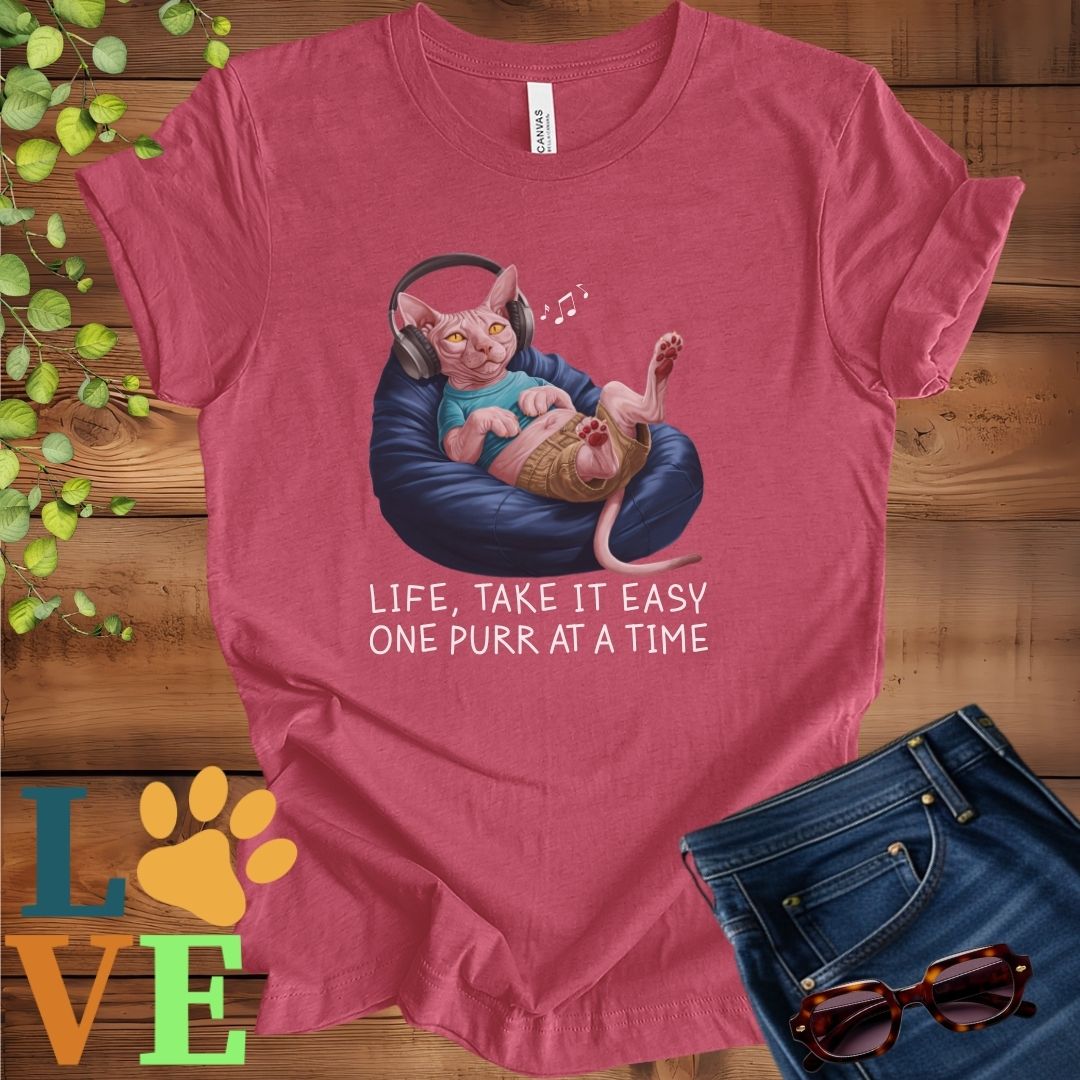 Muted raspberry pink (heather raspberry) t-shirt featuring an anthropomorphic Sphynx cat relaxing on a bean bag while listening to music through headphones, with the slogan "Life, take it easy. One purr at a time."