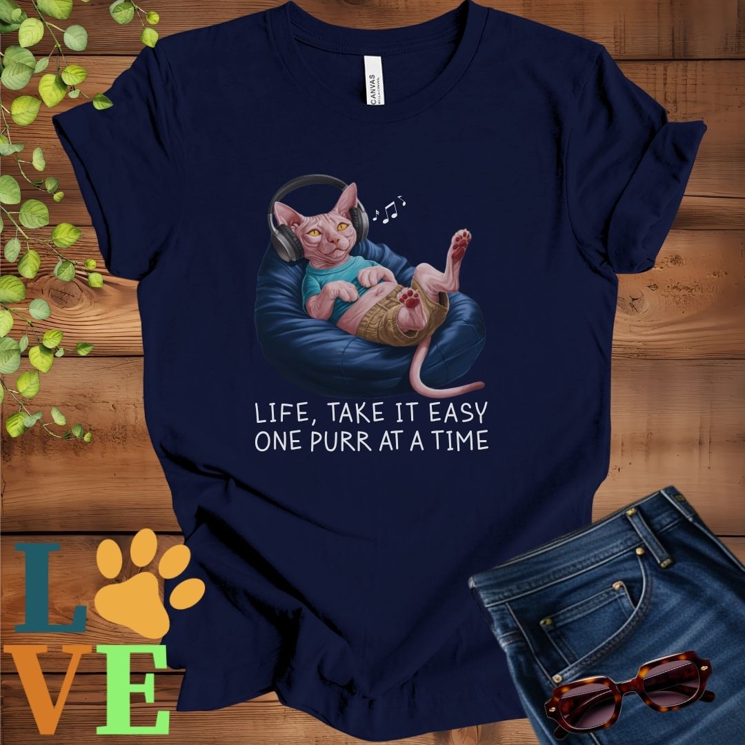 Navy blue t-shirt featuring an anthropomorphic Sphynx cat relaxing on a bean bag while listening to music through headphones, with the slogan "Life, take it easy. One purr at a time."