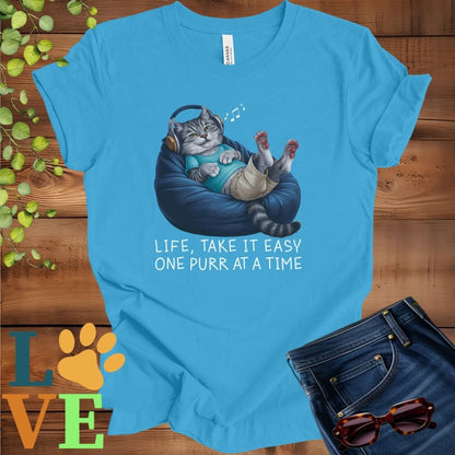 Bright aqua blue t-shirt featuring an anthropomorphic tabby cat relaxing on a bean bag while listening to music through headphones, with the slogan "Life, take it easy. One purr at a time."